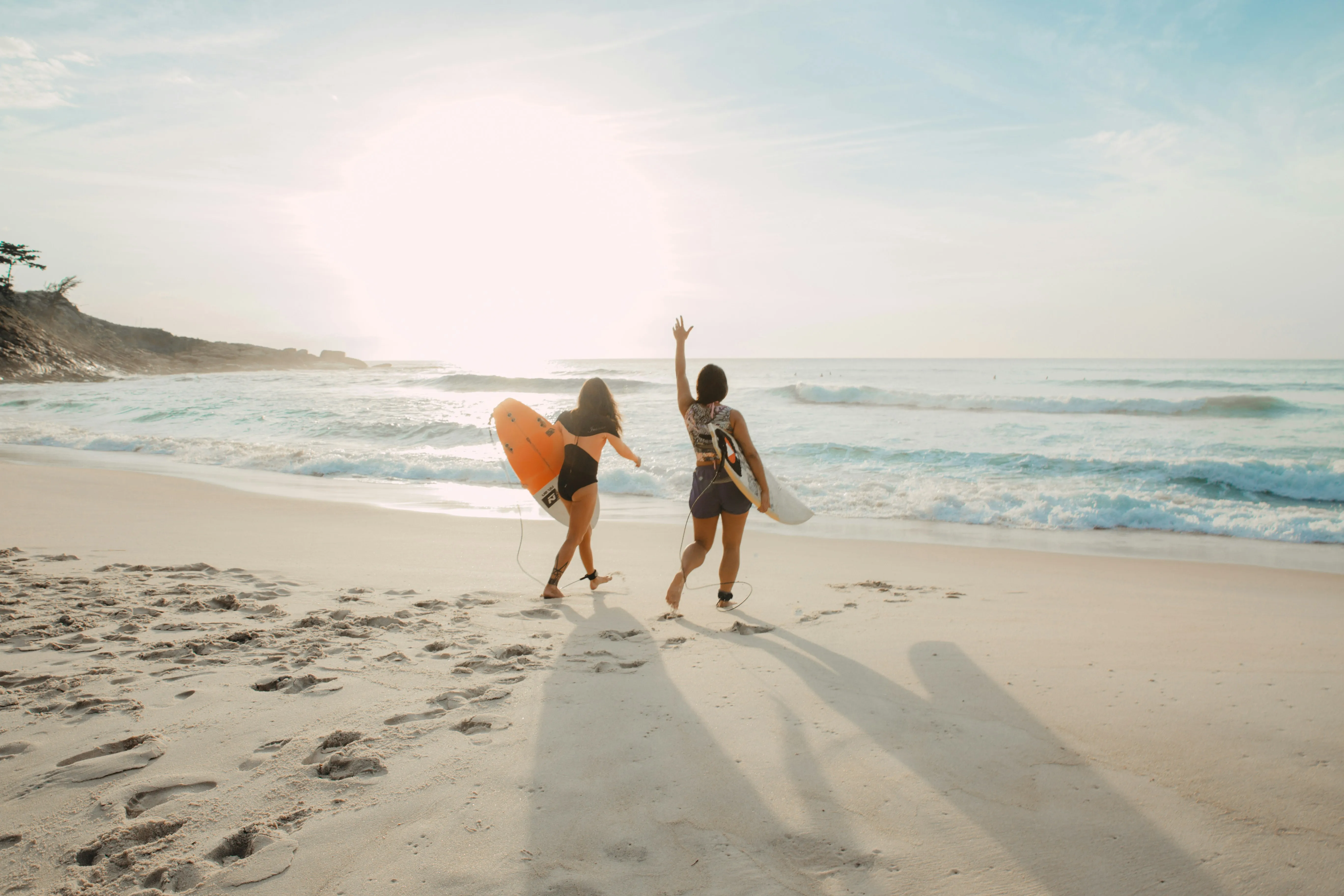 Zwei Surferinnen mit Surfboard am Strand. Die eine winkt in Richtung Meer.