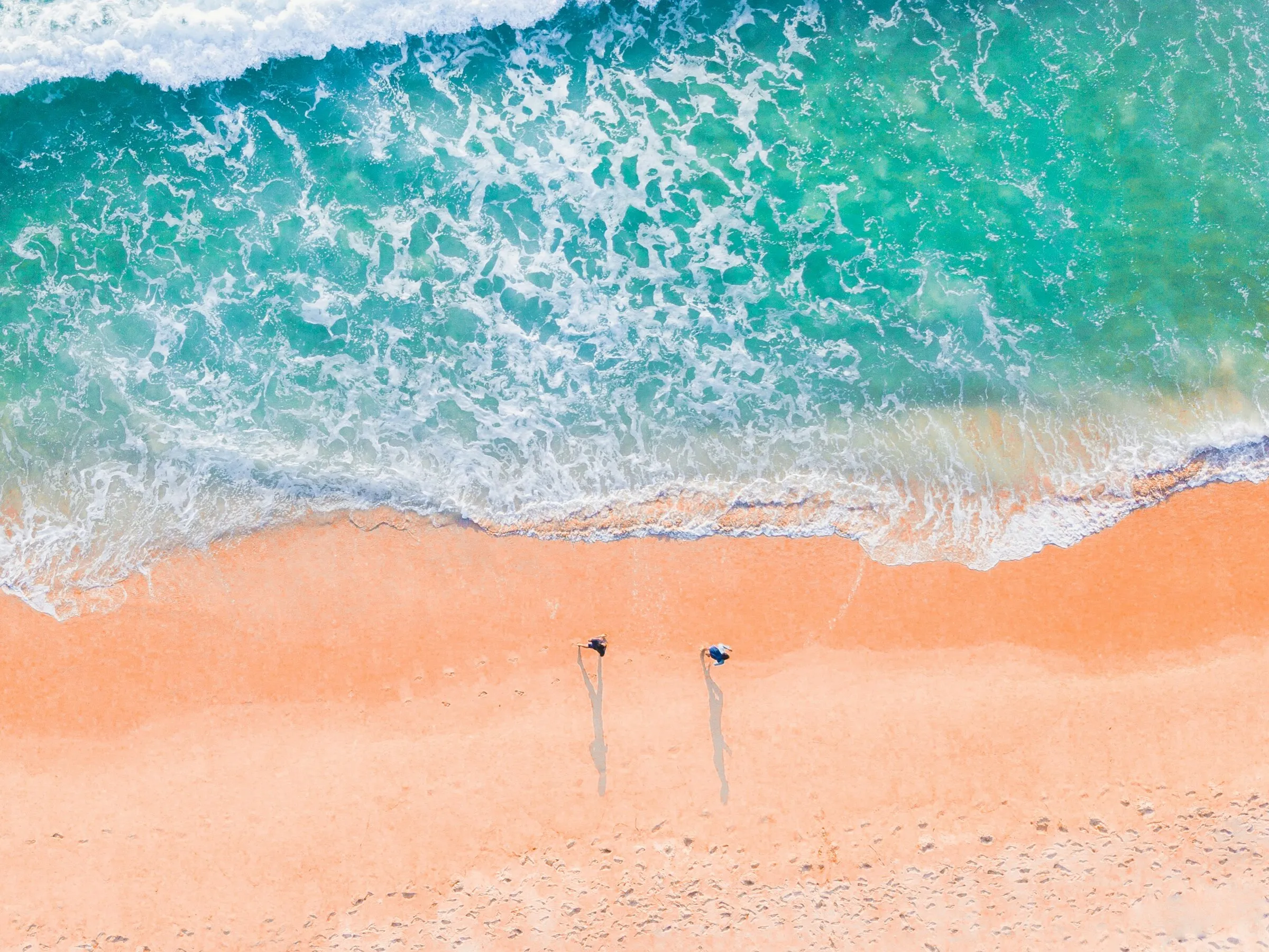 Zwei Personen laufen am Strand im Sand und man sieht sie aus der Vogelperspektive