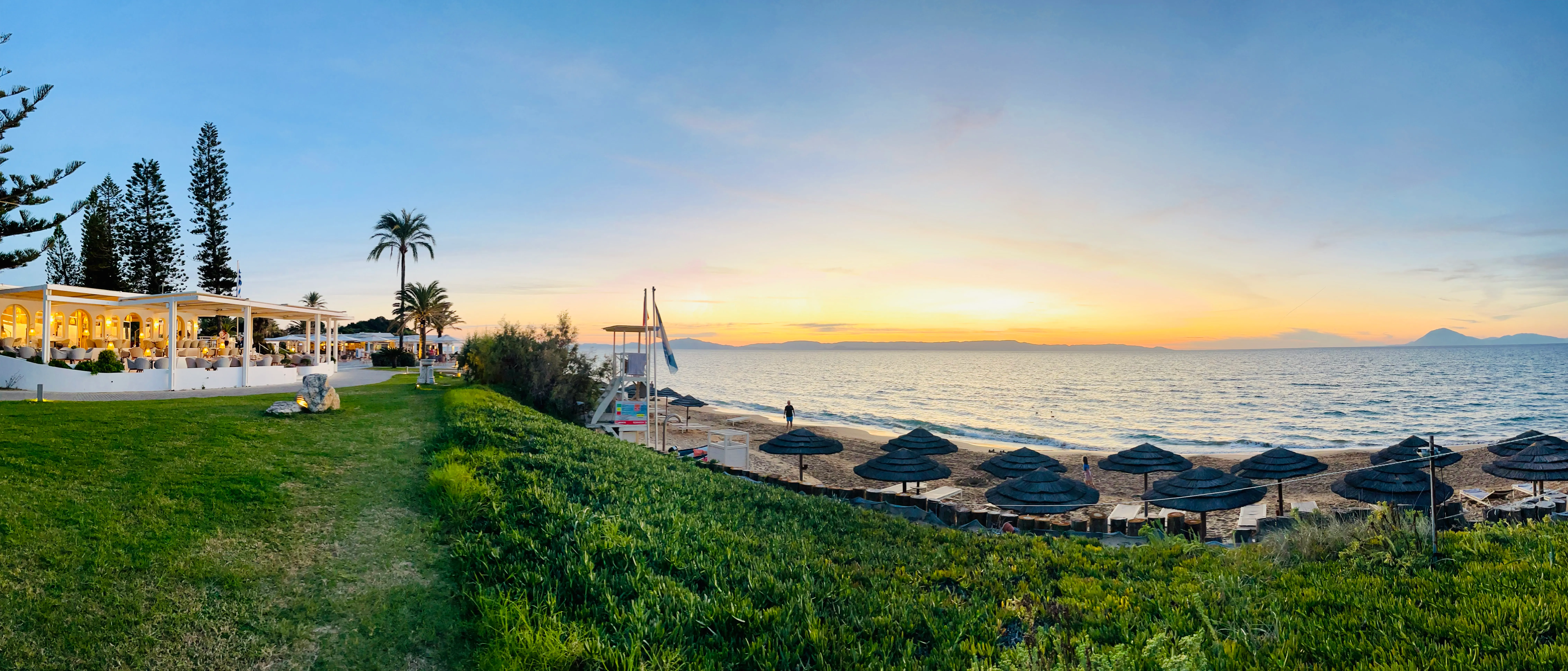 Zwei Surferinnen mit Surfboard am Strand. Die eine winkt in Richtung Meer.
