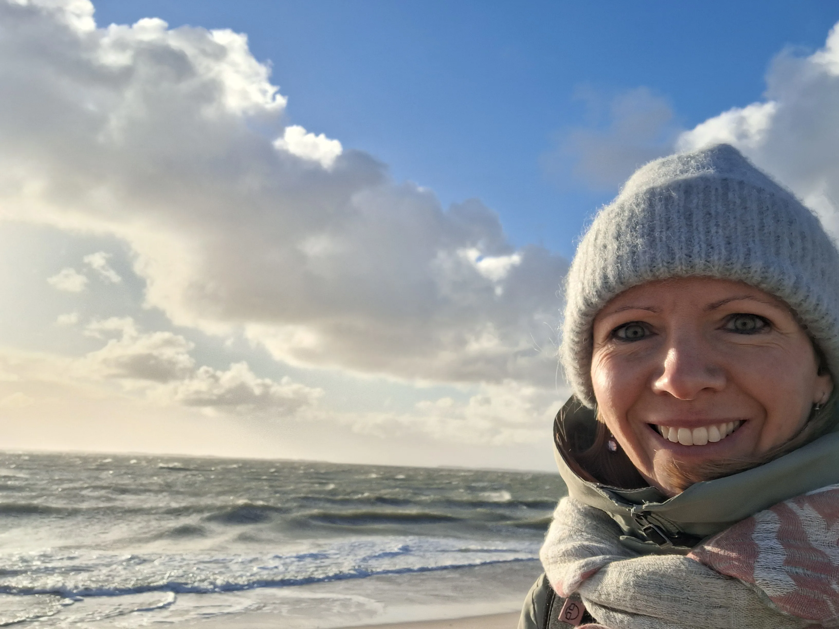 Frau mit Mütze, dicker Jacke und Schal lächelt in die Kamera und im Hintergrund ist die raue See mit ein paar Wolken am Himmel zu sehen