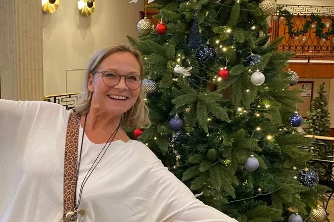 Frau mit weißem Oberteil und Leopardenmuster-Umhängetasche vor einem geschmückten Weihnachtsbaum mit Kugeln und Lichterketten in einem Innenraum