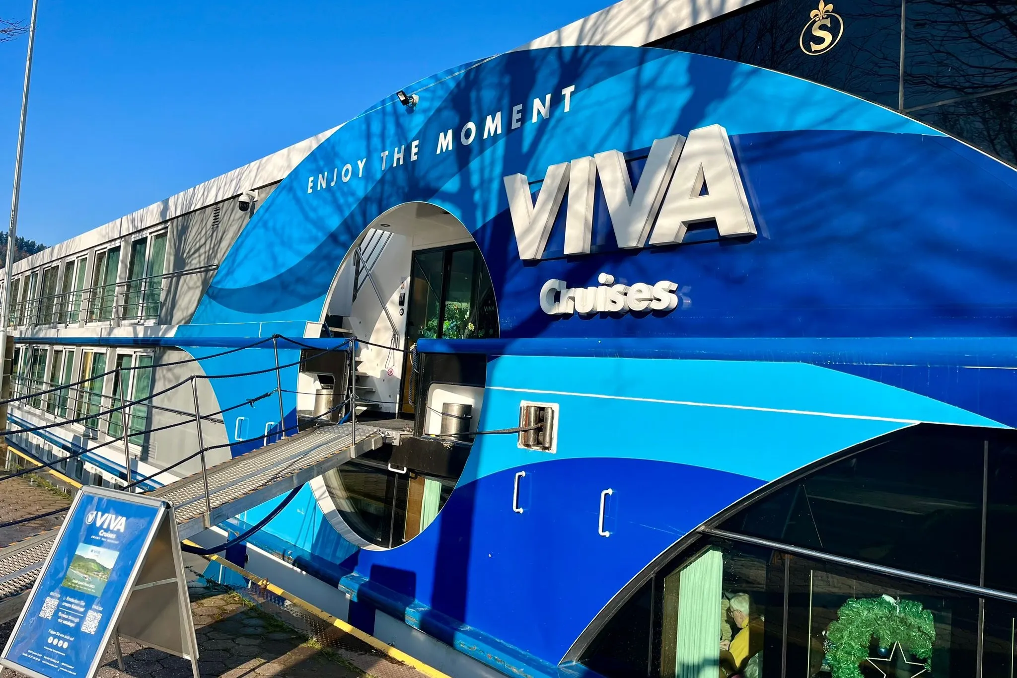 Eingang eines Flusskreuzfahrtschiffes mit der Aufschrift 'ENJOY THE MOMENT VIVA Cruises' und einem Aufsteller davor