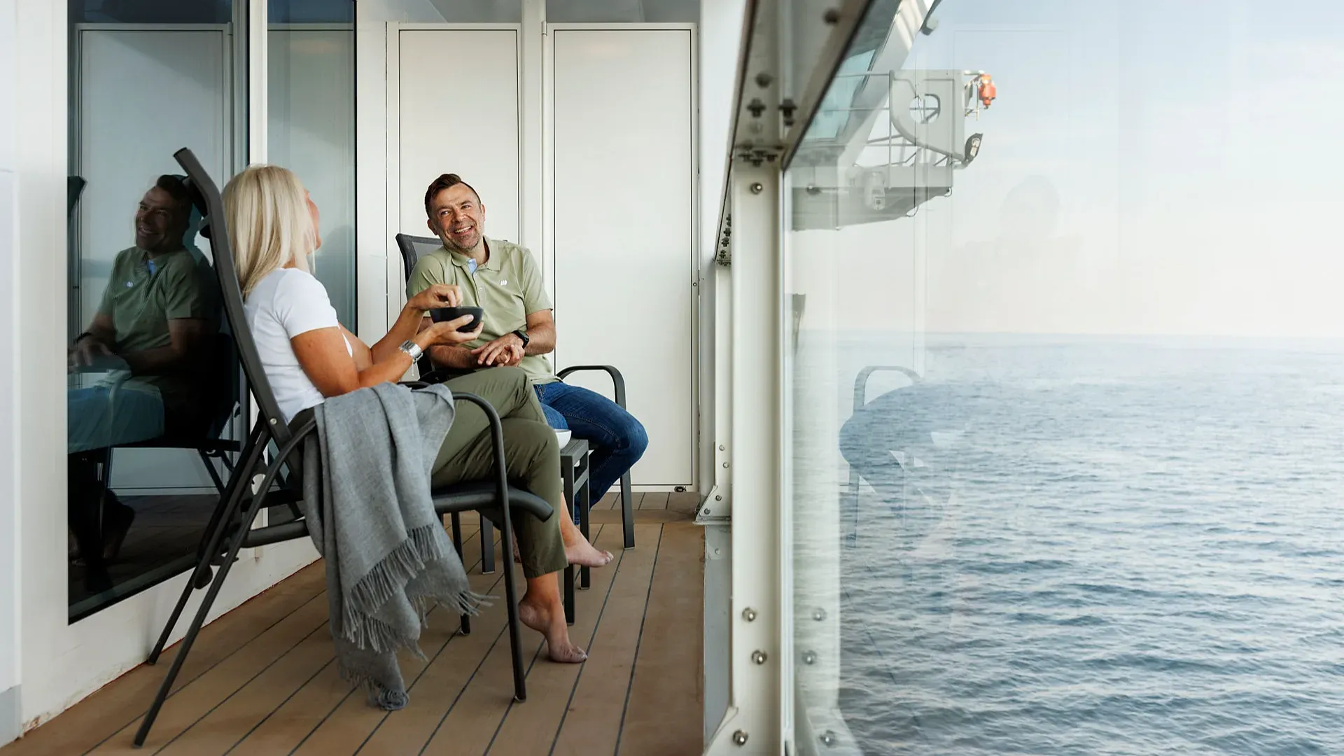 Paar steht auf dem Deck eines Kreuzfahrtschiffs und blickt aufs Meer, Frau hebt den Arm