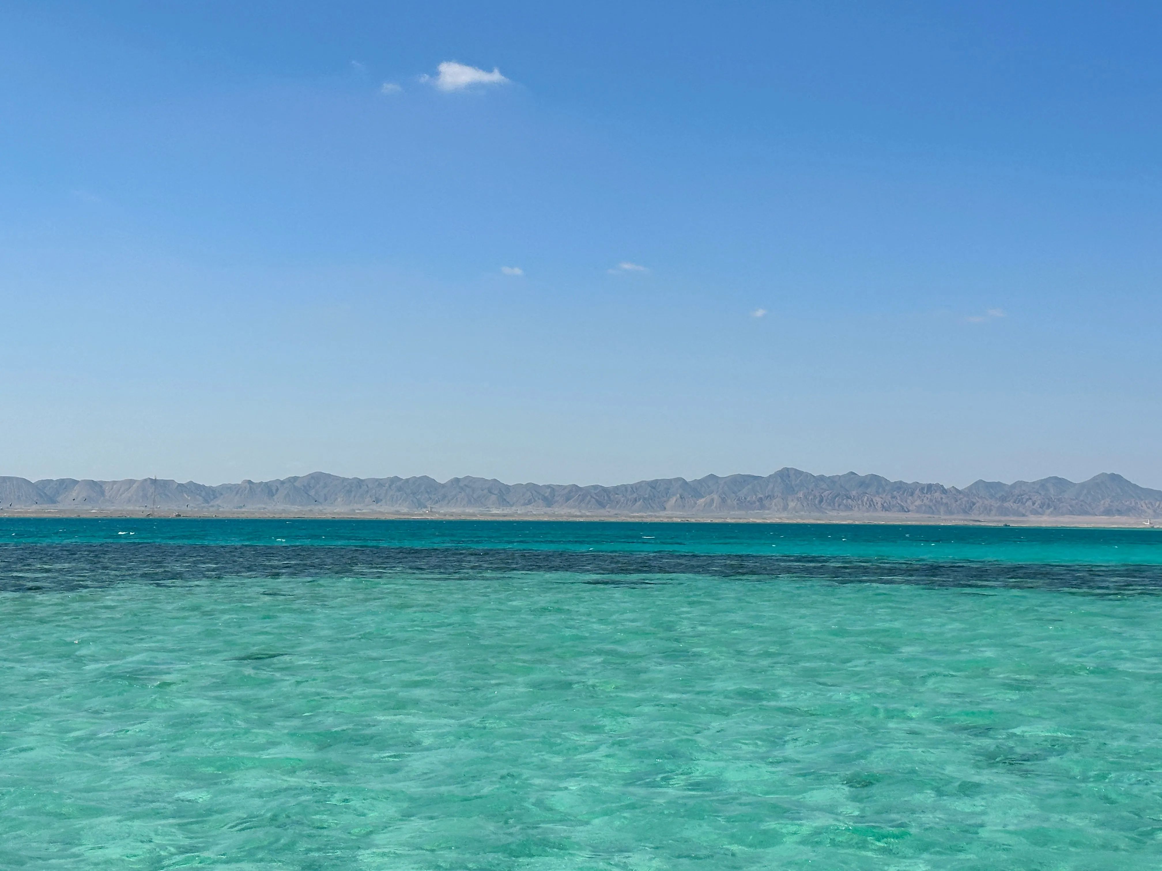 Klares türkisfarbenes Meer mit flachem Wasser vor einer fernen Gebirgskette unter blauem Himmel mit wenigen Wolken