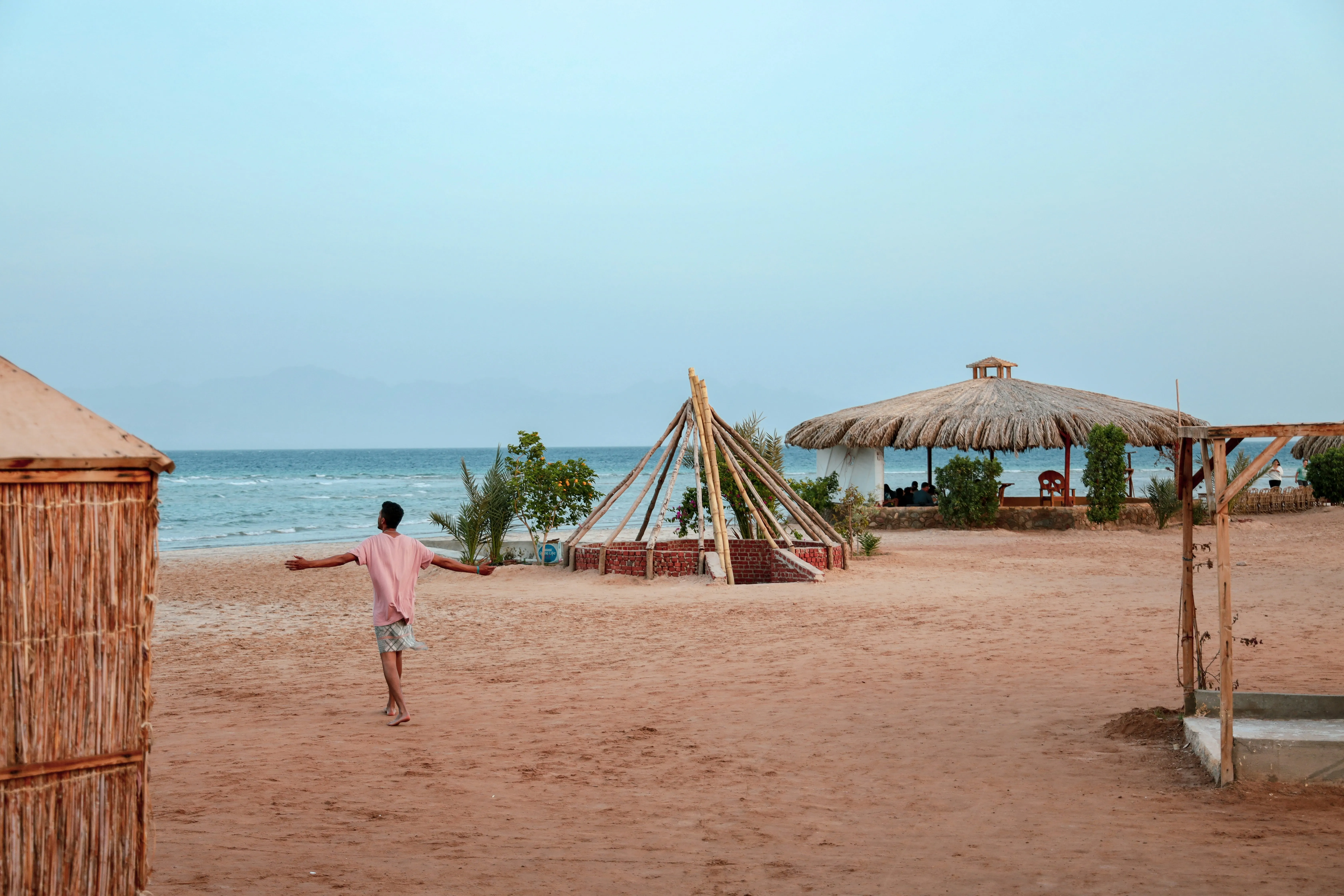 Mann in rosa T-Shirt und Shorts läuft barfuß am Sandstrand mit Strohhütten und Meer im Hintergrund