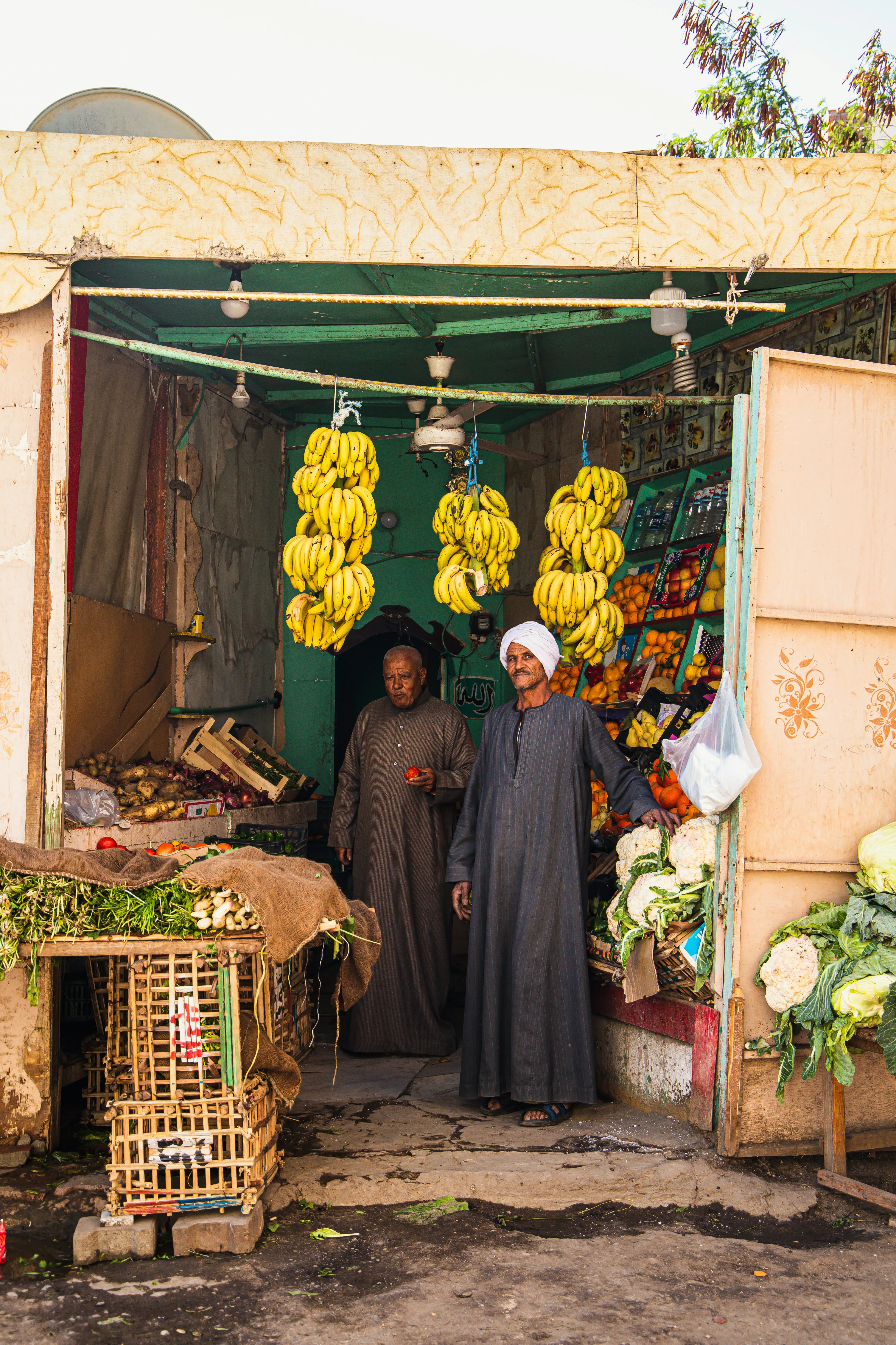 Zwei Personen in dunkler traditioneller Kleidung stehen in einem kleinen Obst- und Gemüsestand mit hängenden Bananen und verschiedenen Früchten und Gemüse auf Regalen