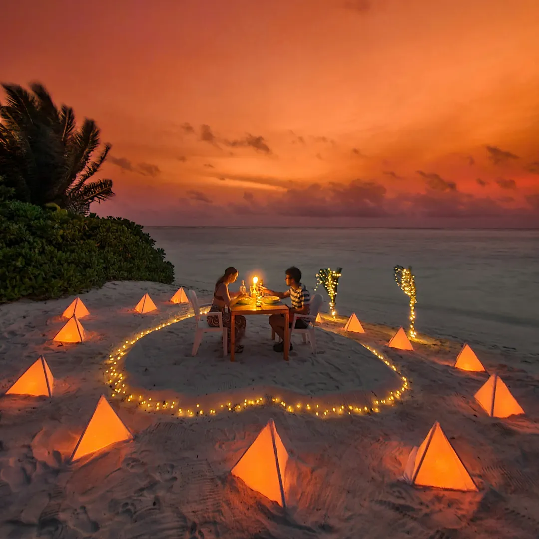 Zwei Personen sitzen an einem kleinen Tisch auf einem Sandstrand, umgeben von leuchtenden Pyramiden und Lichterketten bei Sonnenuntergang
