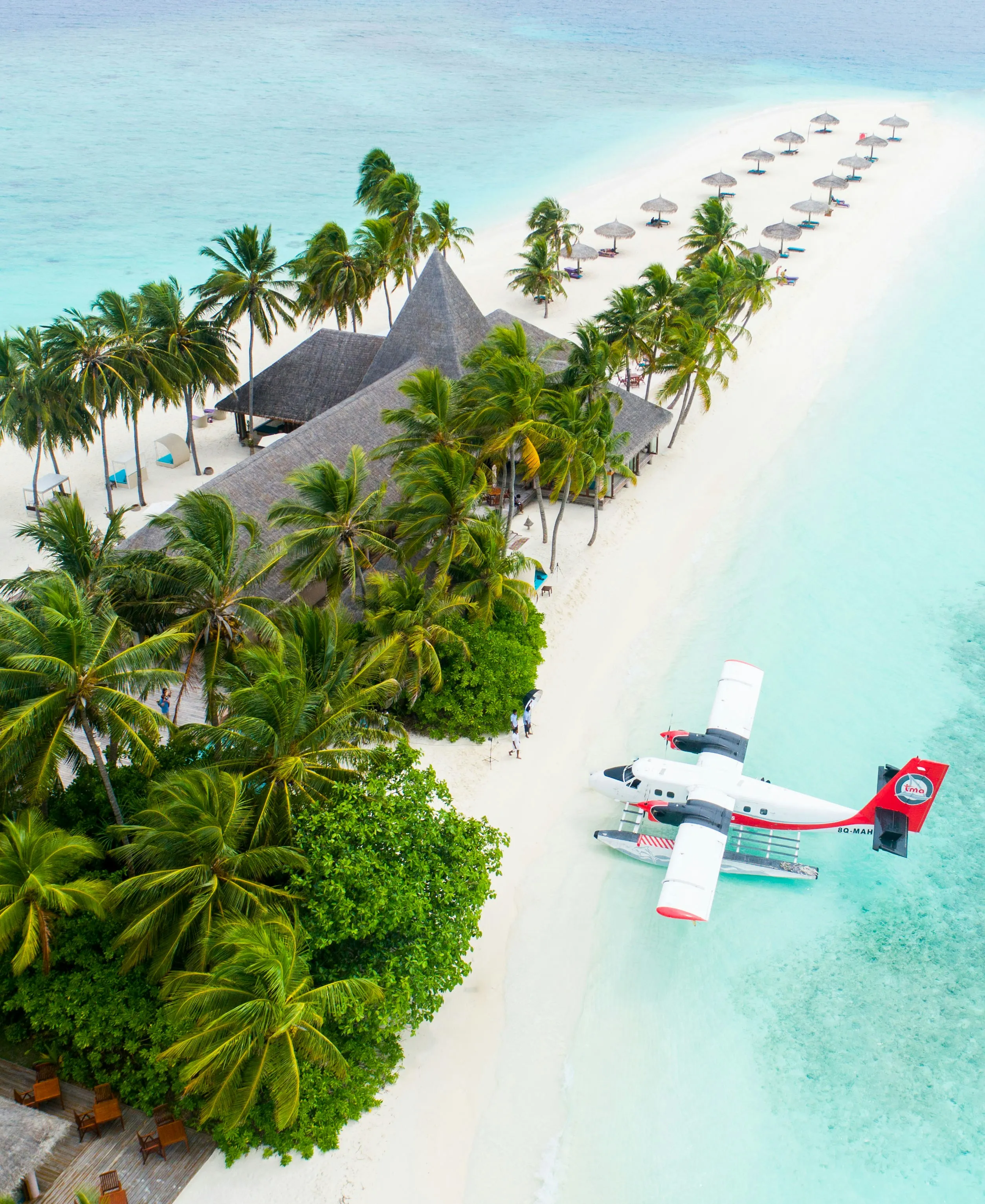 Luftaufnahme einer tropischen Insel mit Palmen, einem strohgedeckten Gebäude, einer Reihe von Sonnenschirmen am Strand und einem Wasserflugzeug im flachen Wasser